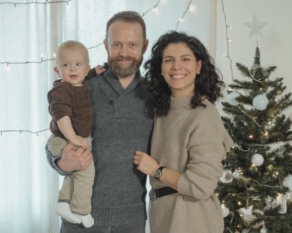 A joyful family with a baby poses next to a decorated Christmas tree indoors.